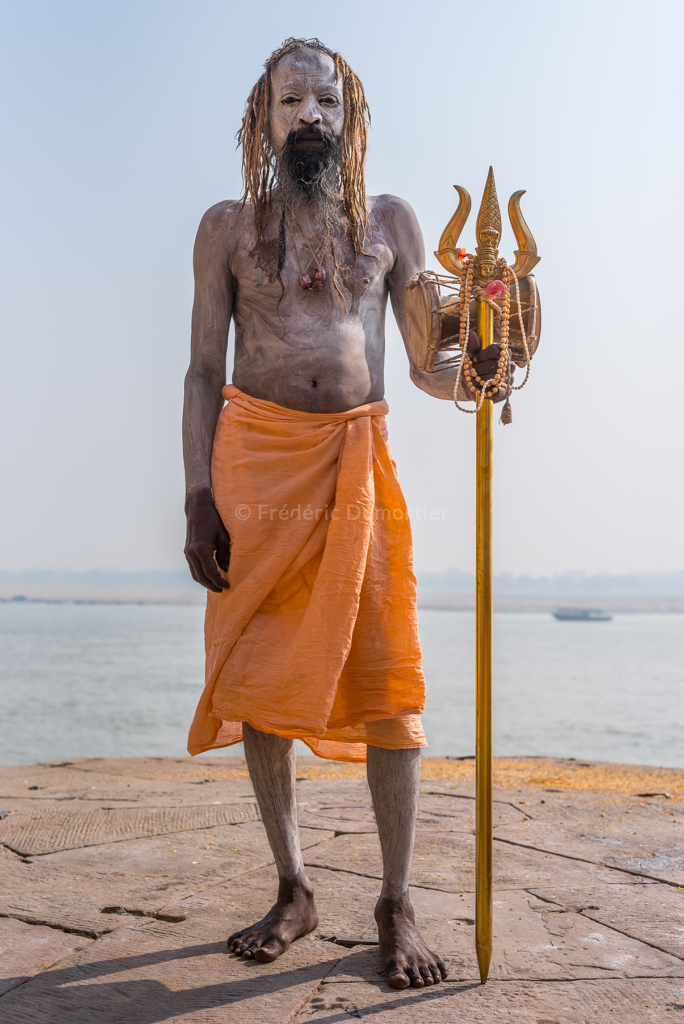 Portrait en pied d'un homme vêtu d'un drap orange noué à la taille, tenant un sceptre recouvert de chapelets. Sa peau est couverte d'une crème blanche. Il se tient au bord de l'eau.