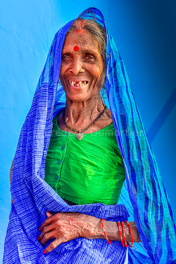 Portrait d'une dame âgée qui sourit face à l'objectif. Elle porte un grand voile bleu vif sur la tête qui retombe sur ses épaules et sa taille. 