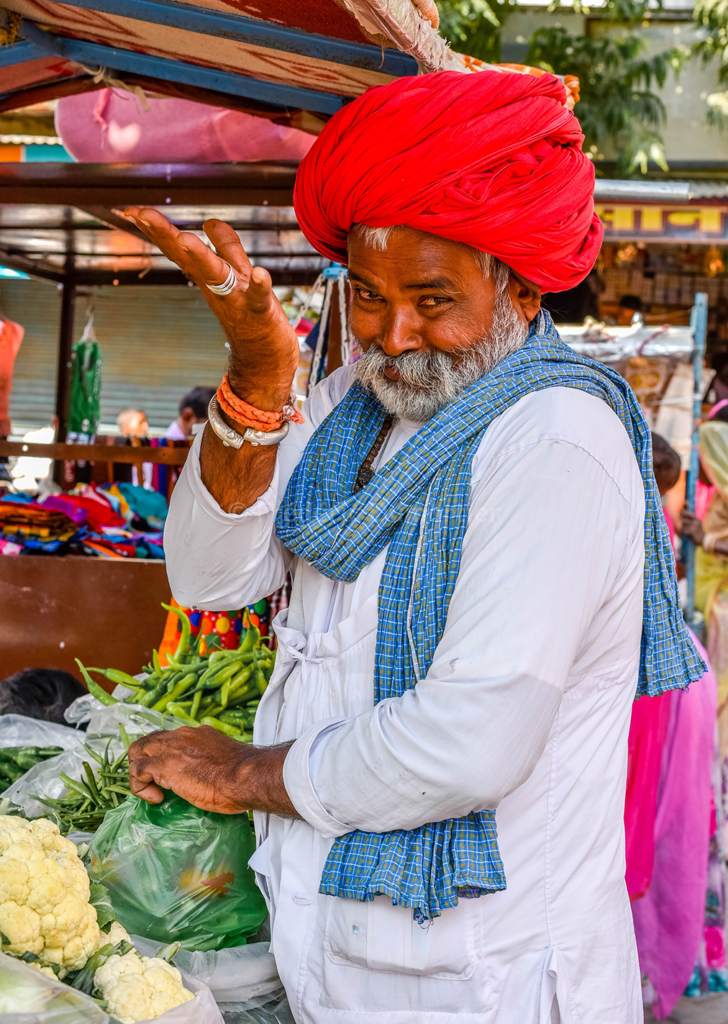 Portrait d'un homme portant un grand turban rouge. Il regarde l'objectif en souriant d'un air malicieux. Il se tient devant l'étal d'un primeur au marché.